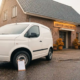 Une camionnette de livraison blanche garée devant un immeuble commercial néerlandais, avec un bloc-notes d'achat appuyé contre le pneu avant.