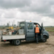 Doi muncitori în veste de lucru urcă într-o dubă încărcată, pe un șantier plin de praf, într-un peisaj industrial olandez plat.
