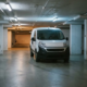 Une camionnette de livraison blanche traverse un parking au plafond bas, aux colonnes de béton étroites et à l'éclairage fluorescent.