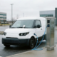 Furgone elettrico bianco collegato a una colonnina di ricarica in un parcheggio commerciale, con la fattura mensile dietro il tergicristallo.