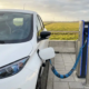 Witte elektrische bestelbus aangesloten op laadpaal bij Nederlandse snelwegservice, groene polderlandschap op achtergrond.