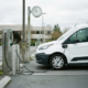Furgonetă electrică albă conectată la o stație de încărcare într-o parcare comercială, ceas analogic pe un stâlp de beton, lumină naturală nordică în condiții de cer înnorat.