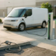 O dubiță electrică albă conectată la o stație de încărcare, cu pompa de combustibil nefolosită pe sol, în lumina caldă a dimineții.