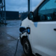 Furgone elettrico bianco collegato al cavo di ricarica al crepuscolo, con l'indicatore della batteria visibile sul cruscotto.
