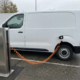 Furgoneta eléctrica blanca conectada a una estación de recarga con un cable de recarga naranja en un aparcamiento comercial de los Países Bajos.