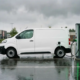 O dubiță electrică albă conectată la un încărcător rapid de curent continuu într-o parcare comercială, cu cablul de încărcare întins între vehicul și stația de încărcare.