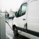 A white electric van plugged into a public charging point in a busy commercial car park in cloudy daylight.