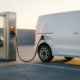 O dubiță electrică albă conectată la o stație de încărcare într-o parcare industrială, în lumina aurie a după-amiezii, cu umbre lungi pe podeaua de beton.