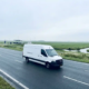 Witte elektrische bestelbus op Nederlandse snelweg met vlak polderlandschap en laadstation op de achtergrond.