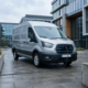 O furgonetă electrică conectată la un cablu de încărcare într-un parc industrial modern, sub cerul înnorat al Europei.