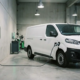 Furgonetă electrică albă încărcată la un încărcător de perete într-un garaj comercial, cablul de încărcare fiind conectat la priza vehiculului.