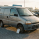 Furgonetă gri-argintiu folosită la un dealer auto, cu lovituri ușoare și un clipboard de inspecție pe anvelopa din față.