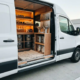 Camionetă albă cu ușă glisantă deschisă și interior organizat cu găleți de vopsea, pensule și mostre de culori în rafturi de perete.